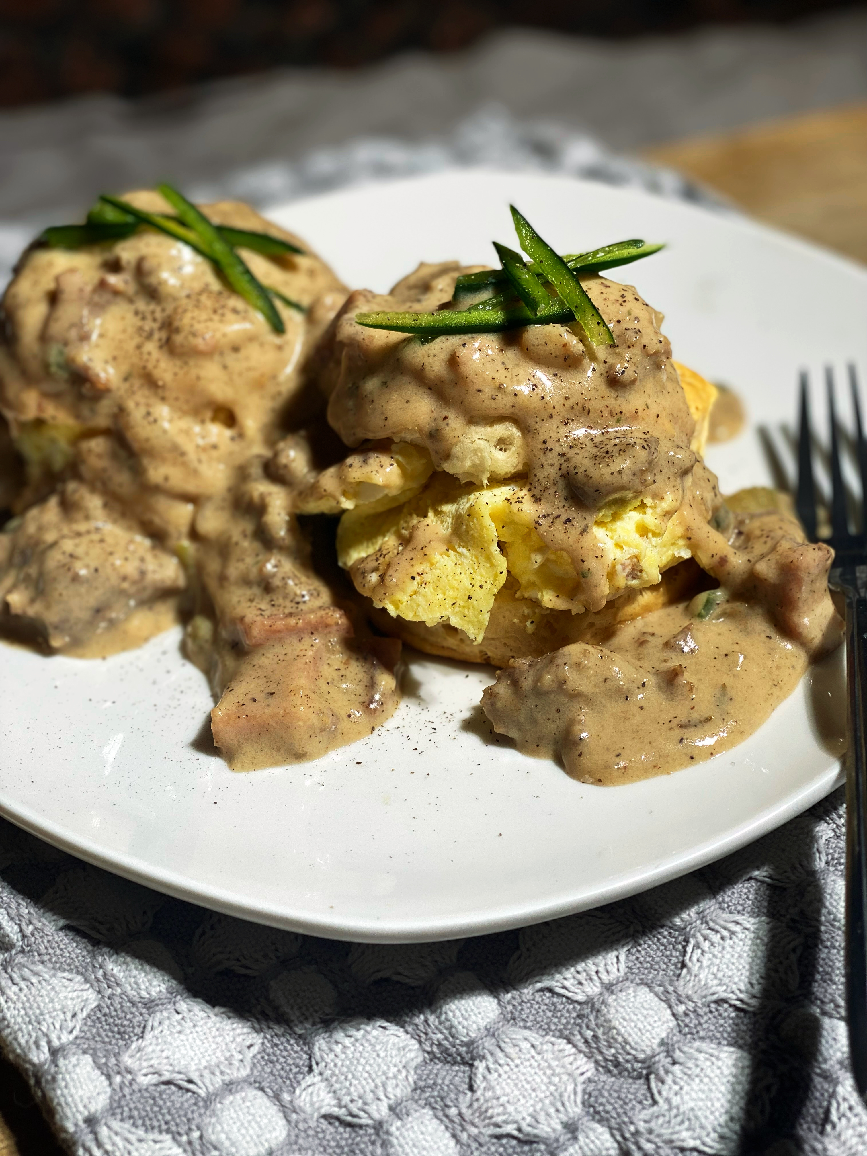 Overhead shot of the plated biscuits and gravy sitting on a grey and white checkered cloth. Biscuit filled with a fluffy scrambled egg, and topped with sausage and ham gravy.